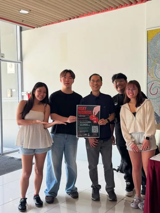 Team behind 'The Hidden Cost' posing with a 'Risky Business' poster